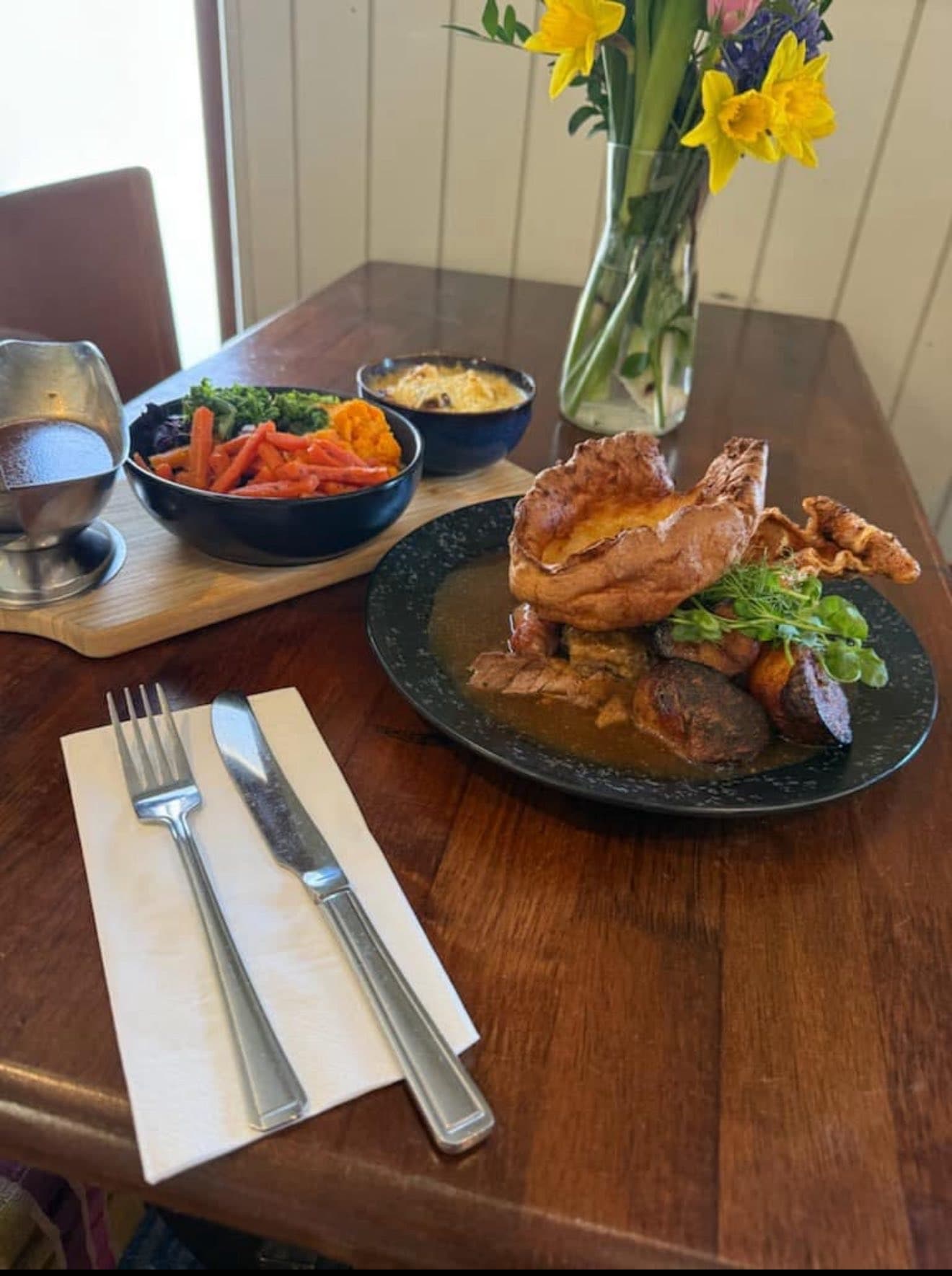 Sunday roast at The George — Yorkshire pudding, sliced roast meat with rich gravy, seasonal vegetables and cauliflower cheese on a wooden table set with daffodils