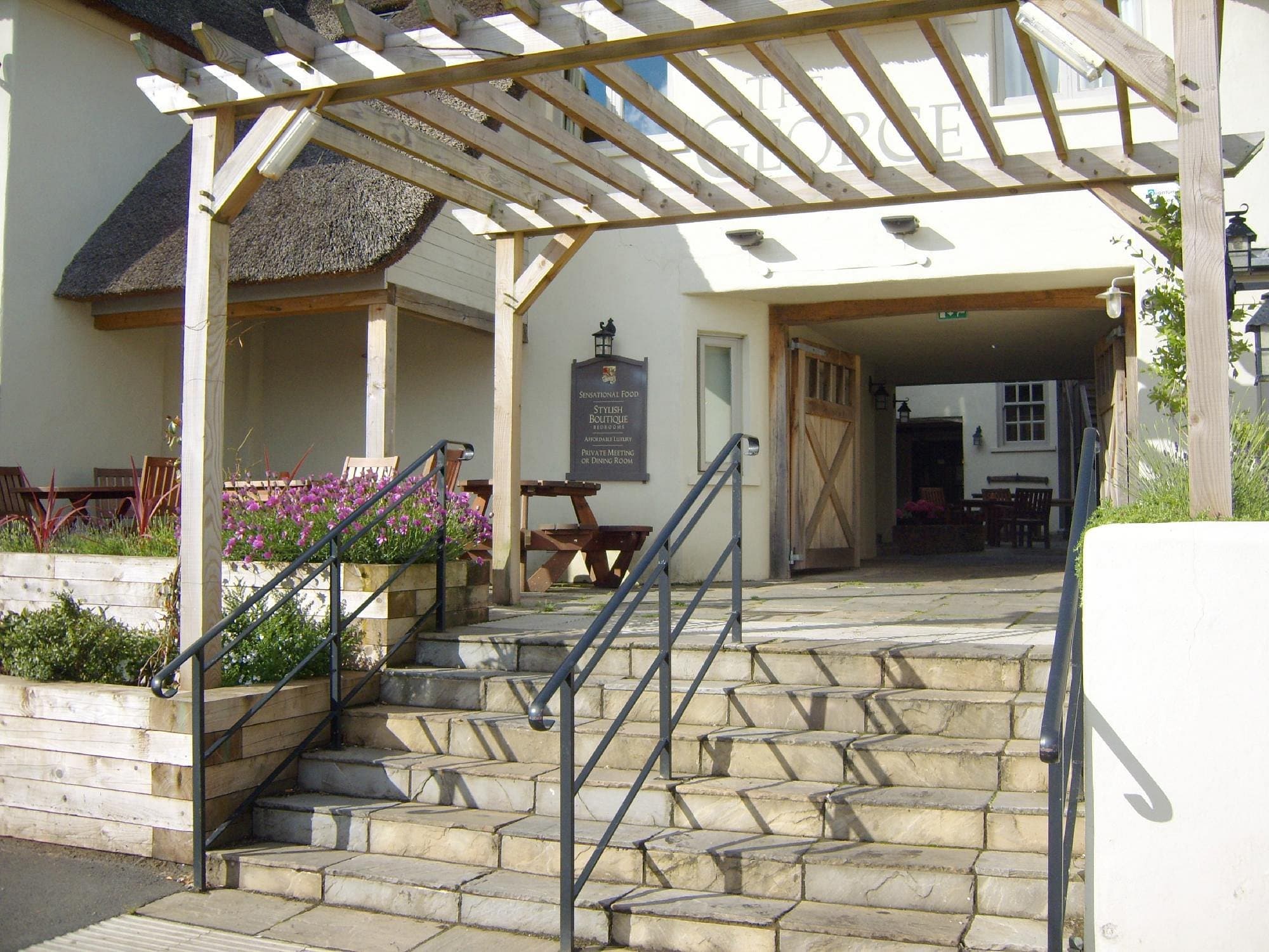 Wooden pergola entrance to The George's courtyard beer garden — picnic tables visible inside, stone steps and summer flowers